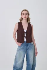 Chaleco sin mangas color marrón oscuro, de corte ajustado al cuerpo, con escote en V y cierre frontal mediante lazos anudados con detalles de cuentas oscuras.