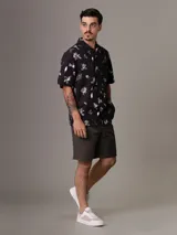Camisa de manga corta con estampado de flores y hojas en blanco y gris sobre fondo negro. Tiene cuello clásico, cierre con botones y bolsillo de parche en el pecho.