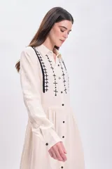 Vestido largo de muselina color crudo con bordado negro en el cuello y la pechera, corte evasé, manga larga y abotonadura frontal.