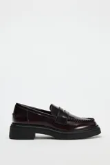 Zapato plano tipo mocasín color borgoña de cuero charol, con detalle de pasados en la parte delantera, copete subido, antifaz y acabado en punta redondeada.