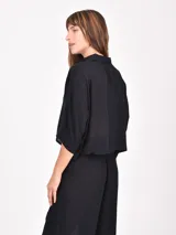 Camisa negra de manga corta con cuello camisero y botones frontales. Presenta un corte cropped y textura acanalada o plisada.