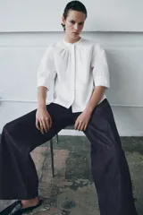 Camisa blanca de algodón con cuello redondo y manga corta abullonada. Presenta un canesú con pliegues verticales en la parte delantera y cierre frontal con botones ocultos. El bajo es redondeado.