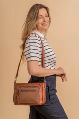 Bandolera de simil cuero color marrón topo, con textura ligeramente desgastada y detalles en nobuck. Presenta múltiples bolsillos frontales con cierres metálicos y tiradores de cuero. Correa ajustable para llevar al hombro o cruzada.