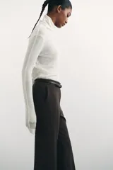 Pantalón de sarga color marrón oscuro, de tiro alto con elástico oculto en la cintura, cierre y pretina prolongada con botón y corchetes ocultos. Tiene piernas con corte para bota, bolsillos al bies, bolsillos de atrás ribeteados y pliegues adelante y atrás.