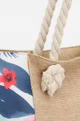 Bolso de playa tipo tote con cuerpo central de tejido rústico color beige (arpillera/yute) y paneles laterales con estampado floral multicolor sobre fondo blanco. Posee asas de cuerda gruesa trenzada.