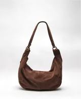 Bolso tipo hobo de gamuza color marrón, con correa de hombro ajustable mediante herrajes metálicos. Presenta un diseño holgado con cierre principal y costuras verticales visibles.