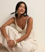 Vestido largo midi color crudo, con escote en V, breteles finos con detalles trenzados, lazo en la espalda, volados y adornos en el bajo.