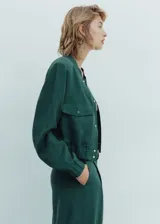 Chaqueta bomber verde esmeralda de lino y lyocell, con cuello redondo, cierre frontal con botones a presión, cintura y puños elásticos, dos bolsillos de parche con solapa en el pecho y corte cropped.
