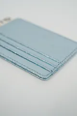 Tarjetero metálico color plateado con textura rugosa y costuras visibles. Incluye una pequeña etiqueta de marca en la parte superior.