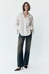 Camisa blanca con rayas verticales grises, de corte holgado, cuello solapa y mangas largas con puños abotonados. Tiene un bolsillo de parche en el pecho, pinzas en la espalda y bajo asimétrico con aberturas laterales.