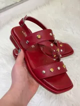 Sandalias rojas de cuero con tiras cruzadas y detalle de tachas plateadas y doradas.