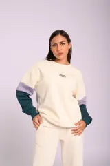 Buzo color crema de felpa con calce oversize, mangas largas con puños en verde oscuro y lila, y estampa bordada con la leyenda "Good Vibes".