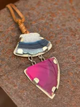 Collar con colgante de dos piedras naturales irregulares engarzadas en metal plateado y dorado, suspendidas de un cordón de cuero marrón con detalle de nudo corredizo. Las piedras tienen vetas en tonos naranja, marrón y blanco.