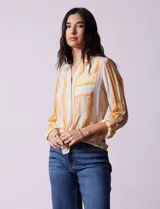 Blusa de gasa estampada a rayas verticales en tonos celeste, lila, naranja y marrón. Tiene cuello a la base con escote en V y botones. Delantera con bolsillo aplicado. Largo de manga regulable con presilla y botón. Terminación anudada opcional.
