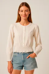 Blusa color blanco con cuello redondo, mangas largas con puños elásticos y cierre frontal con botones.