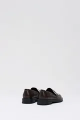 Mocasín tipo penny loafer de eco-piel color marrón chocolate, con suela negra gruesa y detalle de pelo con estampado de vaca en blanco y marrón en el empeine.