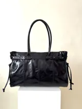 Bolso grande de cuero marrón con textura tipo cocodrilo, estructura rígida y asas para llevar al hombro. Tiene una tira con tachas y hebillas en cada extremo, también lleva lazos en los costados.