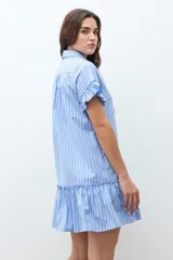 Vestido camisero corto color celeste con rayas blancas y finas rayas rojas. Presenta cuello camisero, botones frontales, mangas cortas con volados y un volado en el ruedo.