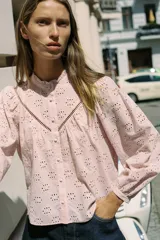 Camisa rosa pálido con bordados perforados, cuello con volante y mangas largas abullonadas. Cierre frontal con botones a tono.