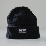 Gorro tipo beanie de tejido de punto acanalado, color negro, con borde doblado y etiqueta rectangular tejida con el logo "PULAU" y el texto "WEAR MORE & WORRY LESS ESTABLISHED IN 2023 IN MONTEVIDEO, UY".