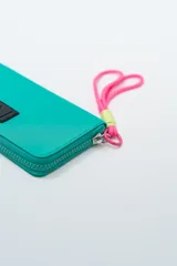Billetera rectangular color fucsia con cierre perimetral y parche frontal negro con logo. Incluye correa de mano de cordón rosa neón y detalle de ajuste en verde lima.
