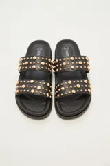 Sandalia tipo slide de color crudo, con dos tiras anchas adornadas con tachas doradas redondas de diferentes tamaños. La suela es gruesa y de goma.