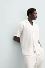 Camisa de calce relajado confeccionada en tejido fluido de mezcla de viscosa, color blanco. Presenta cuello bowling, manga corta, aberturas laterales en el bajo y cierre frontal con botones al tono.