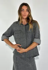 Chaqueta color gris con textura, de corte recto, cuello camisero, cierre frontal con botones, mangas 3/4 con puños arremangados, dos bolsillos de parche en el pecho y ruedo recto.