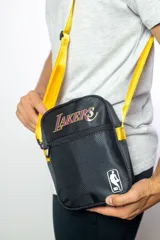 Morral cruzado de nylon negro con textura de rombos, con el logo de Los Angeles Lakers en tonos morado y dorado, y el logo de la NBA en blanco en la parte inferior. Posee correa regulable de color amarillo.