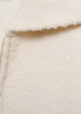 Abrigo corto color crudo de tejido bouclé con mezcla de lana. Tiene cuello de solapa ancha, cierre delantero con cremallera, manga larga con puños elásticos y terminaciones de canalé en contraste. Diseño oversize.