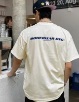 Remera de algodón color beige claro, corte oversize y manga corta. Presenta un estampado en la espalda con el texto "BOHEMIAN DSG®" en azul y debajo "CERTIFIED AUTHENTIC STREETWEAR" en letras más pequeñas.