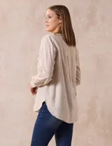 Blusa color beige de lino y rayón, con cuello a la base, escote en V y abertura con botones. Tiene bolsillos en la parte delantera y mangas largas regulables con presilla y botón.
