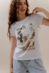 Remera blanca de algodón con cuello redondo y manga corta. Presenta una estampa ilustrada de una mujer con sombrero sentada en un interior, rodeada de plantas y mirando a través de una ventana con vista a palmeras.