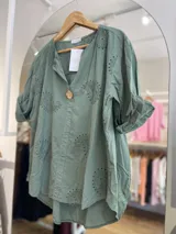 Blusa color crema con cuello camisero, mangas cortas con dobladillo y cierre frontal con un botón de madera. Presenta bordados con motivos de paisley y perforaciones.