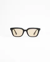 Lentes de sol de montura gruesa color negro, estilo cuadrado/rectangular, con cristales de tono ámbar/beige claro. Presentan detalles de remaches metálicos en las esquinas frontales.