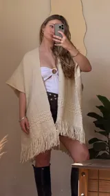 Ruana color crema tejida a mano en lana merino peinada, con diseño calado y flecos en el borde inferior.