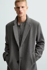 Abrigo straight fit color beige confeccionado en tejido con lana. Cuello con solapas de muesca, manga larga acabada en puño con botones, bolsillos de vivo en cadera, detalle de bolsillo interior y bajo con abertura en centro espalda. Cierre frontal de botonadura.