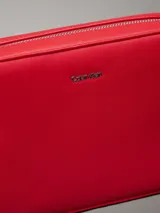 Cartera cruzada tipo 'camera bag' de Calvin Klein, color rojo vibrante, con forma rectangular y esquinas redondeadas. Presenta un acabado liso y el logo metálico de la marca en el centro frontal. Incluye herrajes metálicos plateados y una correa ajustable con detalles en tela.
