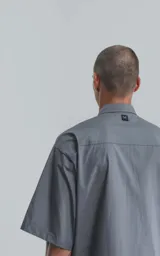Camisa gris de manga corta confeccionada en poplin 100% algodón, con silueta oversize y dos bolsillos delanteros, uno con solapa y otro con ojal y botón.