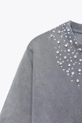 Camiseta gris de algodón con efecto lavado, cuello redondo y mangas cortas. Decoración con apliques de strass plateados en el escote.