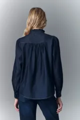 Camisa de denim azul oscuro con cuello mao, corte recto, mangas largas con puños abotonados, cierre frontal con botones a presión y detalle de volados en el cuello y la pechera.