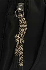 Bolso tipo tote de nylon negro, con asas de mano y correa ajustable para hombro. Presenta un bolsillo frontal con solapa y un detalle de cordón trenzado colgante.