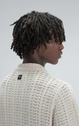 Camisa de tejido de punto color beige, manga corta, con cuello polo y cierre frontal de botones. Presenta un calce regular y un patrón calado en zigzag.