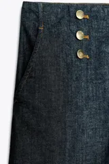 Pantalón culotte de tiro alto en denim azul oscuro, con pernera ancha y corta terminada en vuelta. Presenta falsos botones dorados en la parte delantera, bolsillos frontales y cierre con cremallera y gancho.