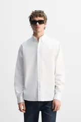 Camisa blanca de corte regular fit confeccionada en popelín de algodón. Tiene cuello abotonado, manga larga con puños abotonados y cierre frontal con botones.