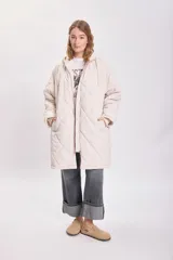 Campera larga color crudo con capucha y cierre frontal, diseño acolchado con patrón de rombos.