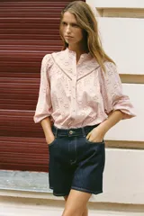 Camisa rosa pálido con bordados perforados, cuello con volante y mangas largas abullonadas. Cierre frontal con botones a tono.