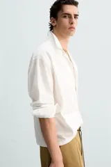 Camisa blanca de corte relajado confeccionada en tejido de algodón y lino. Tiene cuello bowling, manga larga con puños abotonados, jaretas en el delantero, bolsillos de parche en pecho y cadera, bajo con aberturas laterales y cierre frontal con botones.