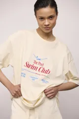 Remera color crema con estampado frontal con la frase "The Peel Swim Club" en letras rojas y azules, y un dibujo de una persona nadando. Corte holgado y mangas cortas.
