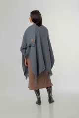 Ruana de punto color gris con diseño de trama diagonal y terminación de flecos en el ruedo. Presenta un pequeño parche decorativo en la parte frontal inferior.